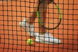 Joueur de tennis portant des tennis blanches tenant une raquette de tennis avec une balle de tennis jaune posée près de ses pieds, derrière le filet du terrain de tennis.
