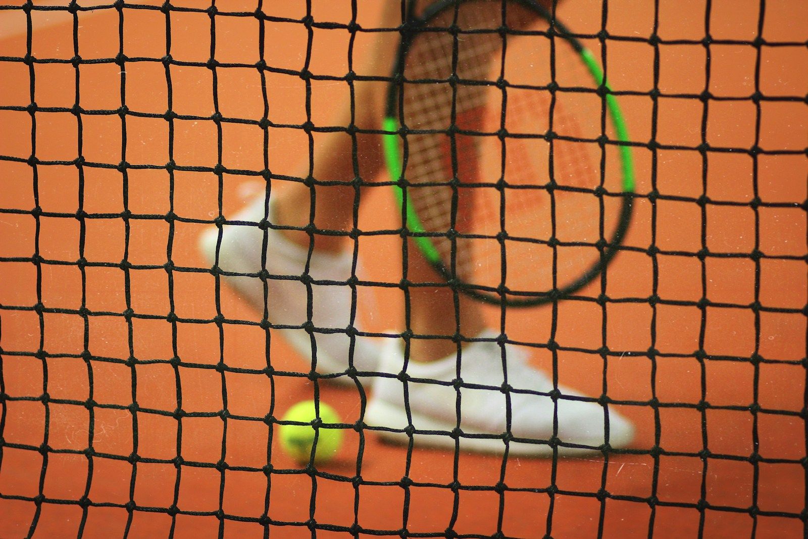 Joueur de tennis portant des tennis blanches tenant une raquette de tennis avec une balle de tennis jaune posée près de ses pieds, derrière le filet du terrain de tennis.
